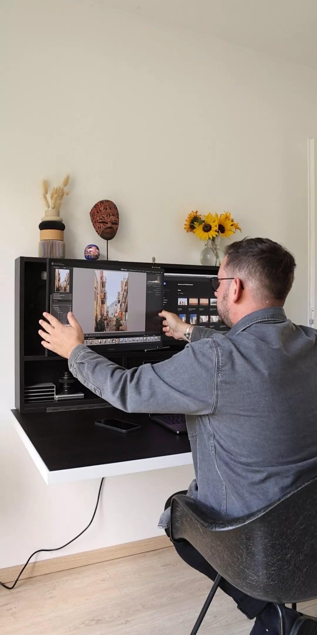 Man sitting at an open black DropTop desk editing photographs on a large monitor, with dried flowers and sunflowers on the shelf above.