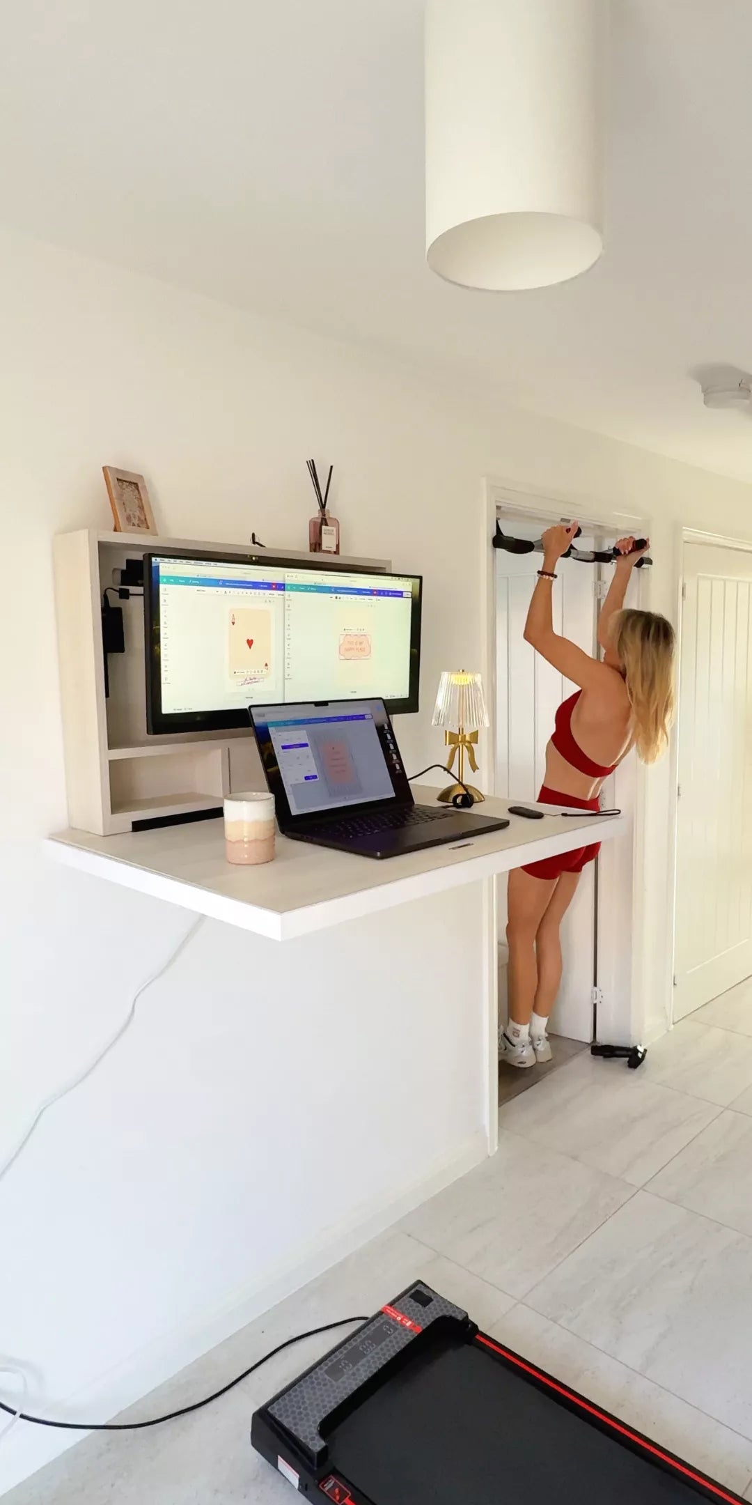 Woman in red sportswear exercising on a pull-up bar beside an open white DropTop desk, with a walking pad on the floor.