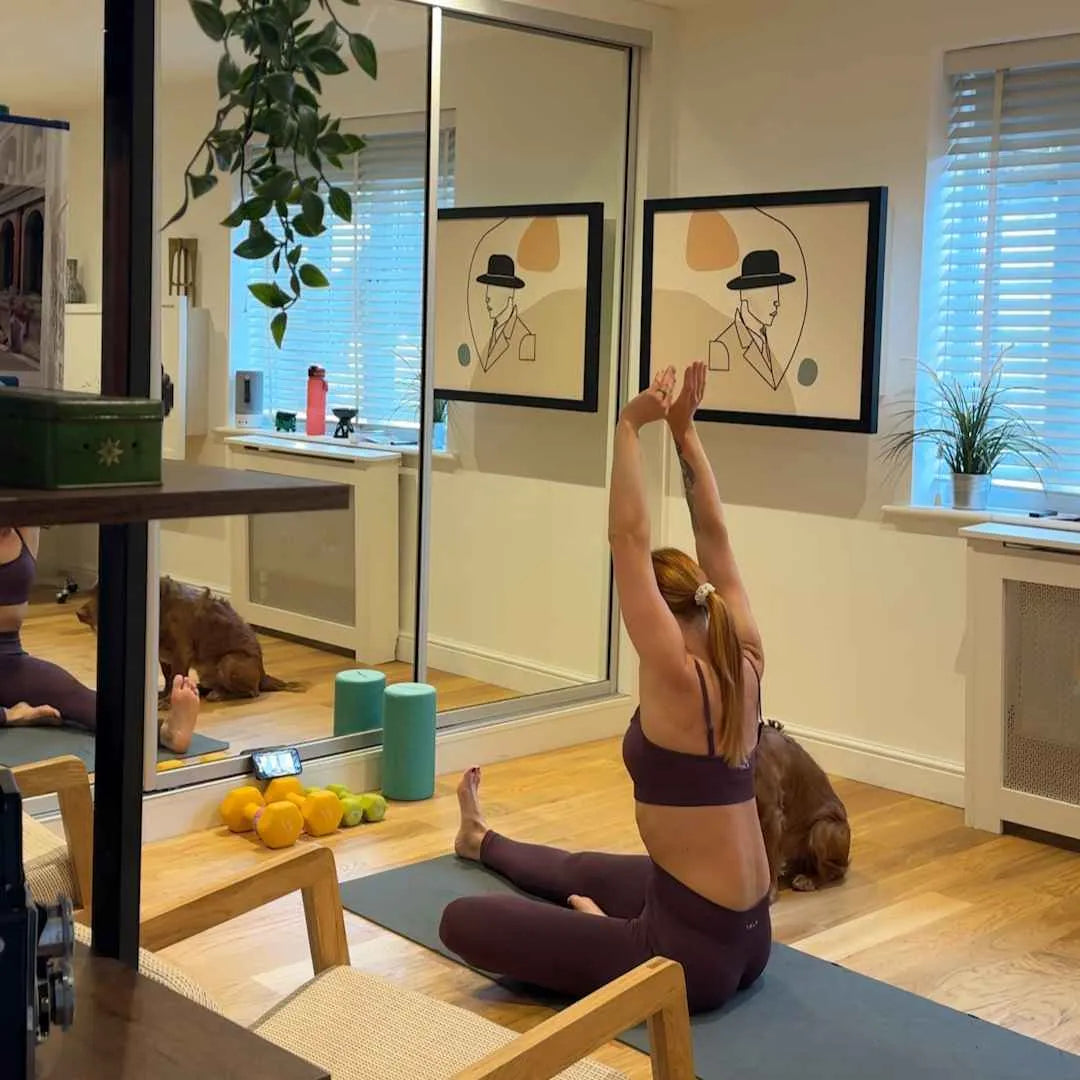 Woman stretching on a yoga mat at home with her dog beside her.
